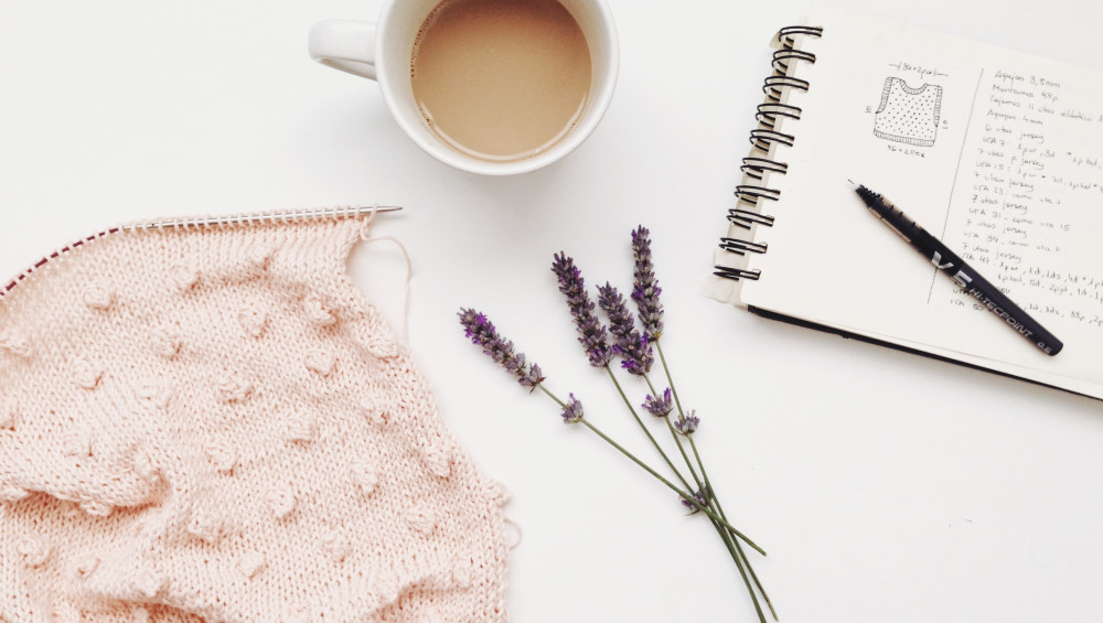 Photo of a knitted vest on a knitting needle, a sprig of lavender, a cup of tea, and a notebook with a knitting pattern written in it.
